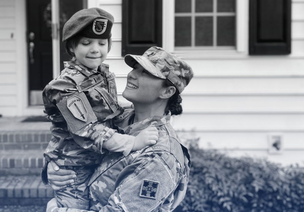A women soldier in uniform holding her son dressed in a mini uniform.