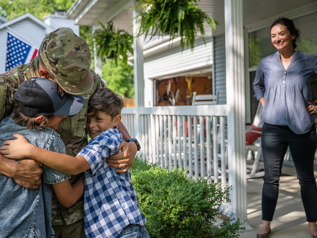 Military spouse watching her husband in uniform hugging their kids