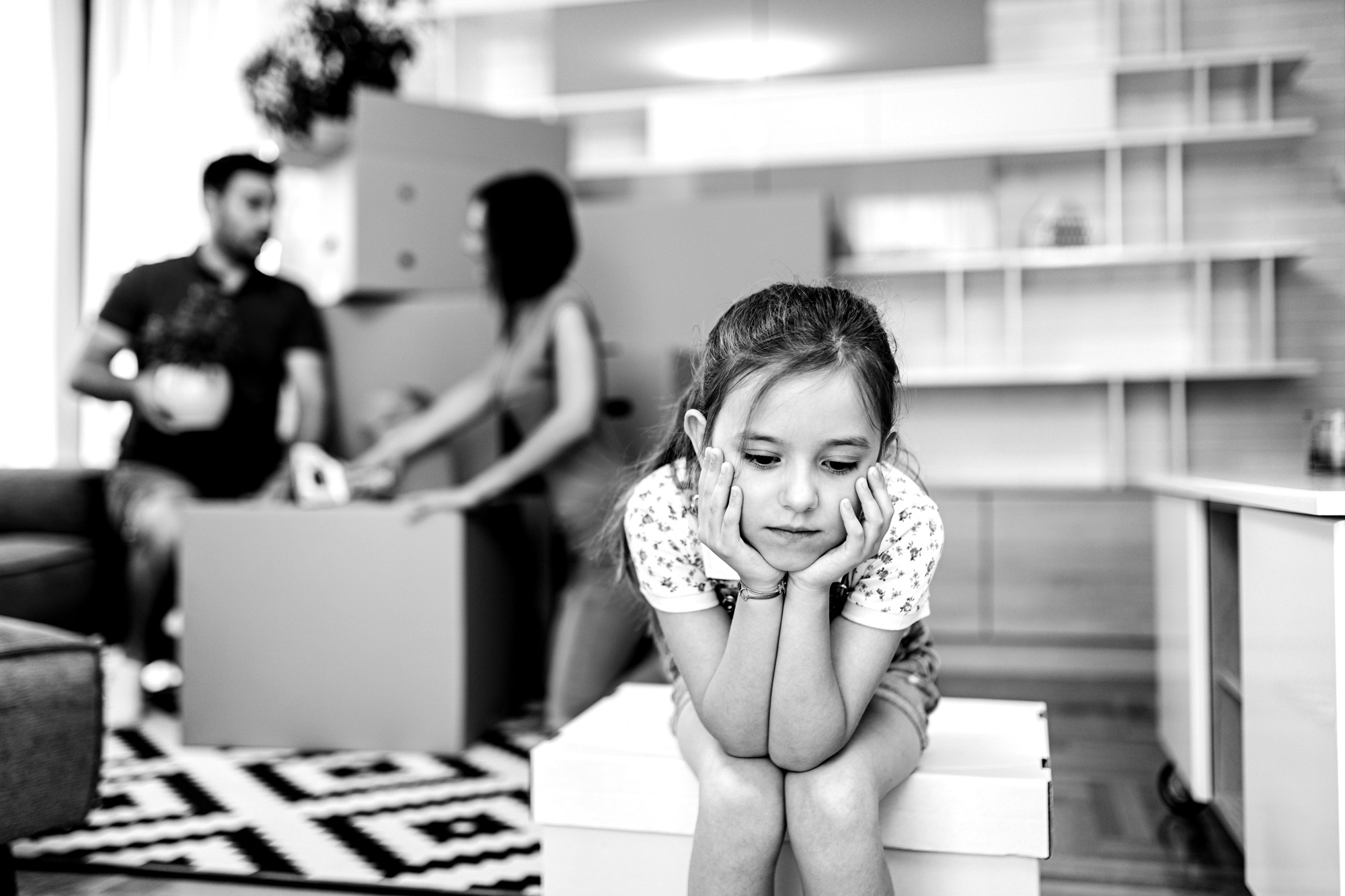 Child sitting alone and upset as two parents are packing boxes to move.
